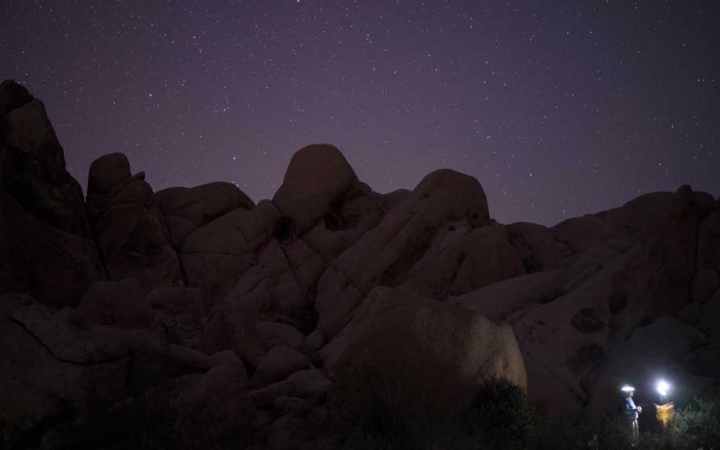 Two people wearing headlamps appear in the bottom right of the photo. Behind them, large boulders are illuminated in front of a purple starry sky.